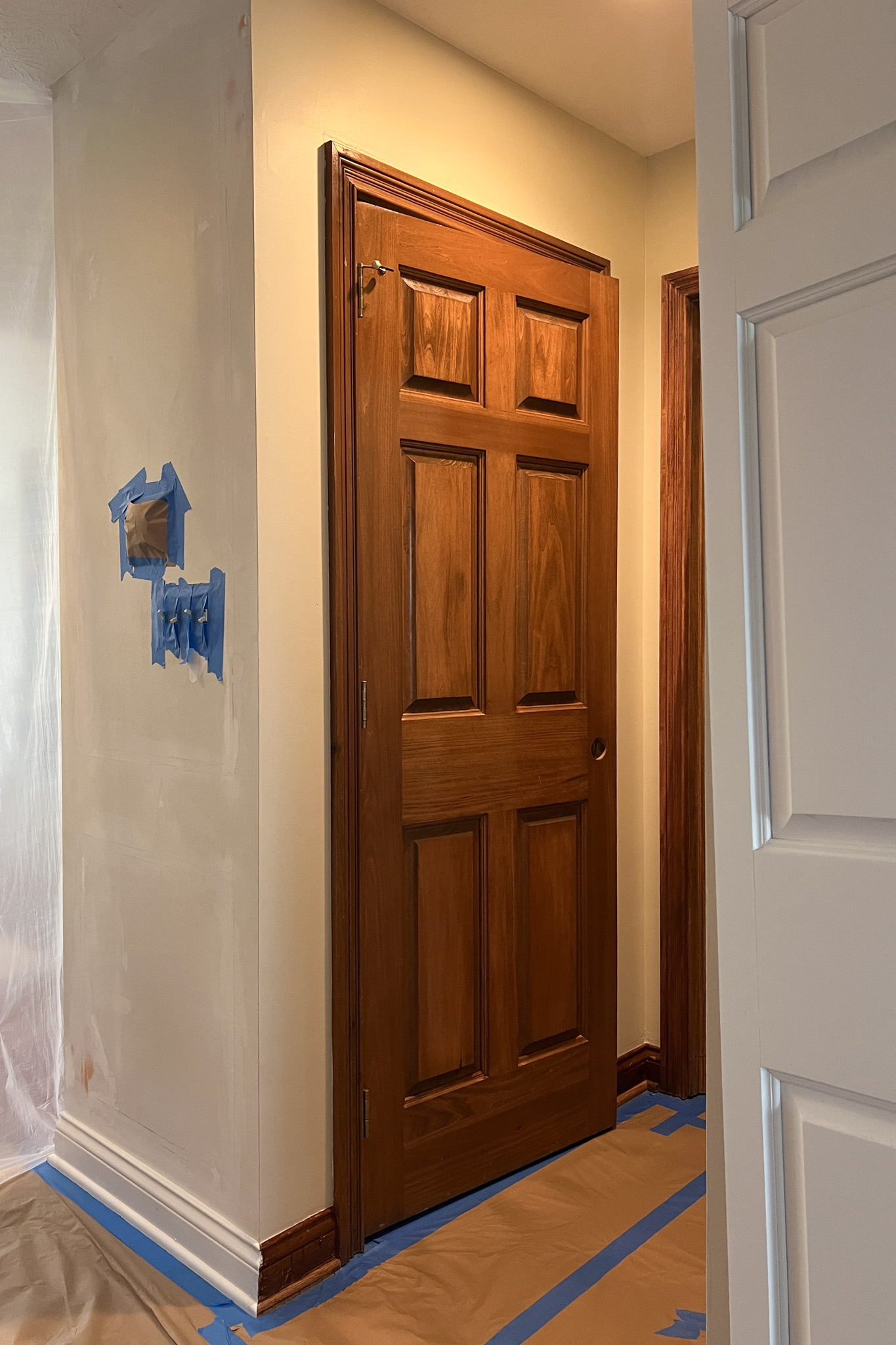 Dark wood six-panel door in an entryway, surrounded by walls prepped for painting with blue tape and drop cloths protecting the trim and floor.