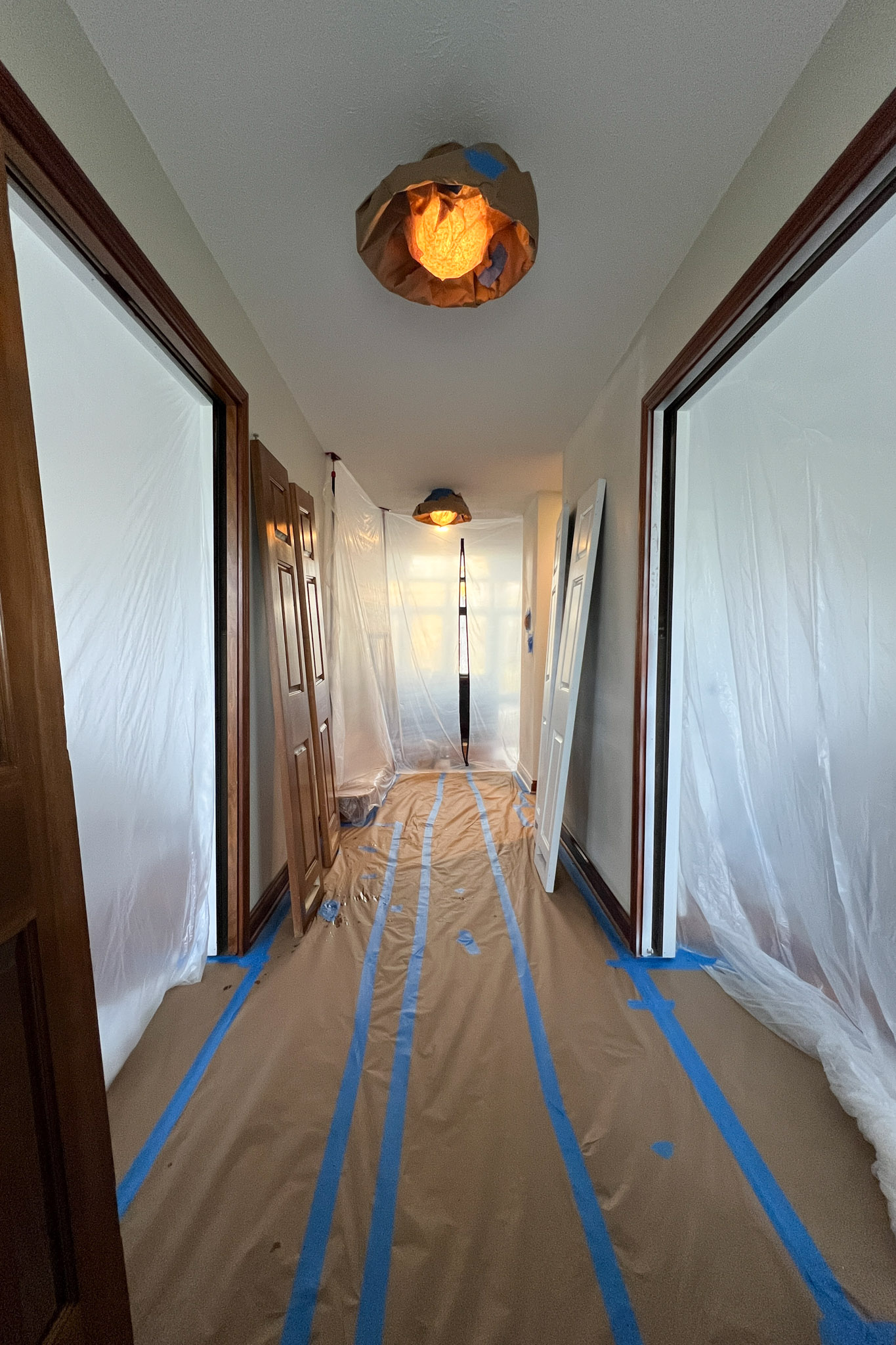 A long hallway prepped for painting with floors covered in paper and tape, plastic sheeting over doorways, and covered light fixtures.