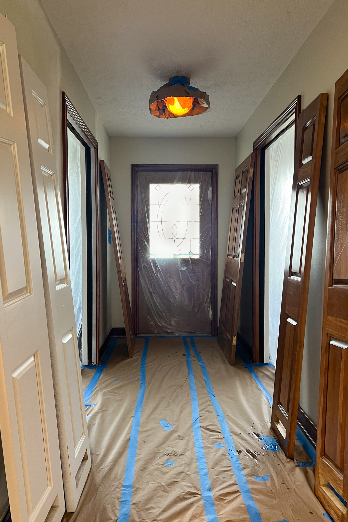 Entryway hallway fully prepped for painting, with the front door covered in plastic and several interior doors leaning against the protected walls and floor.