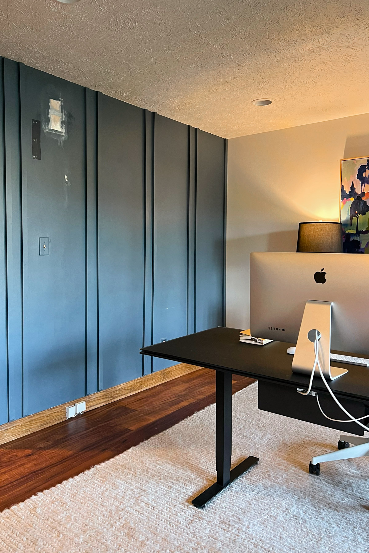  A home office featuring a black standing desk with an iMac, a textured white rug, and a navy blue paneled accent wall.