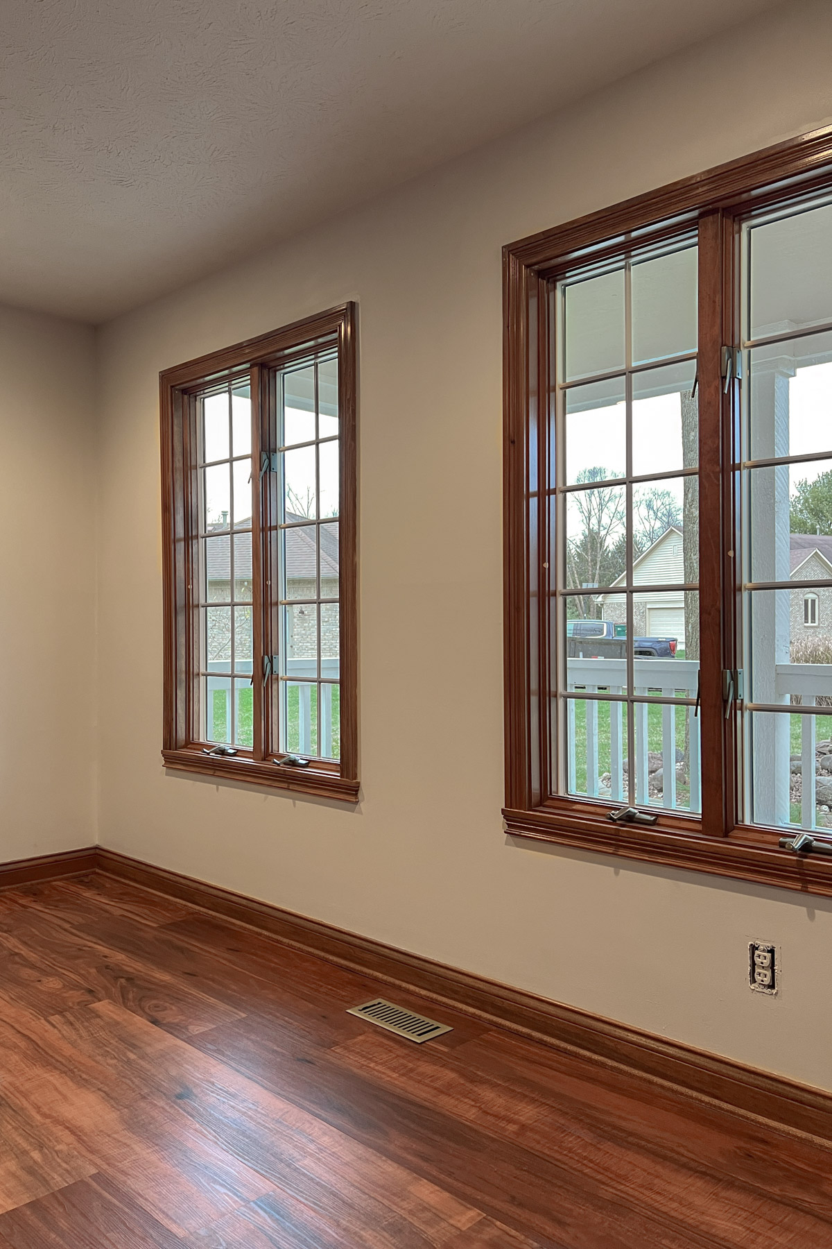 An empty room with polished reddish-brown hardwood floors, light beige walls, and two large casement windows framed in matching wood trim.