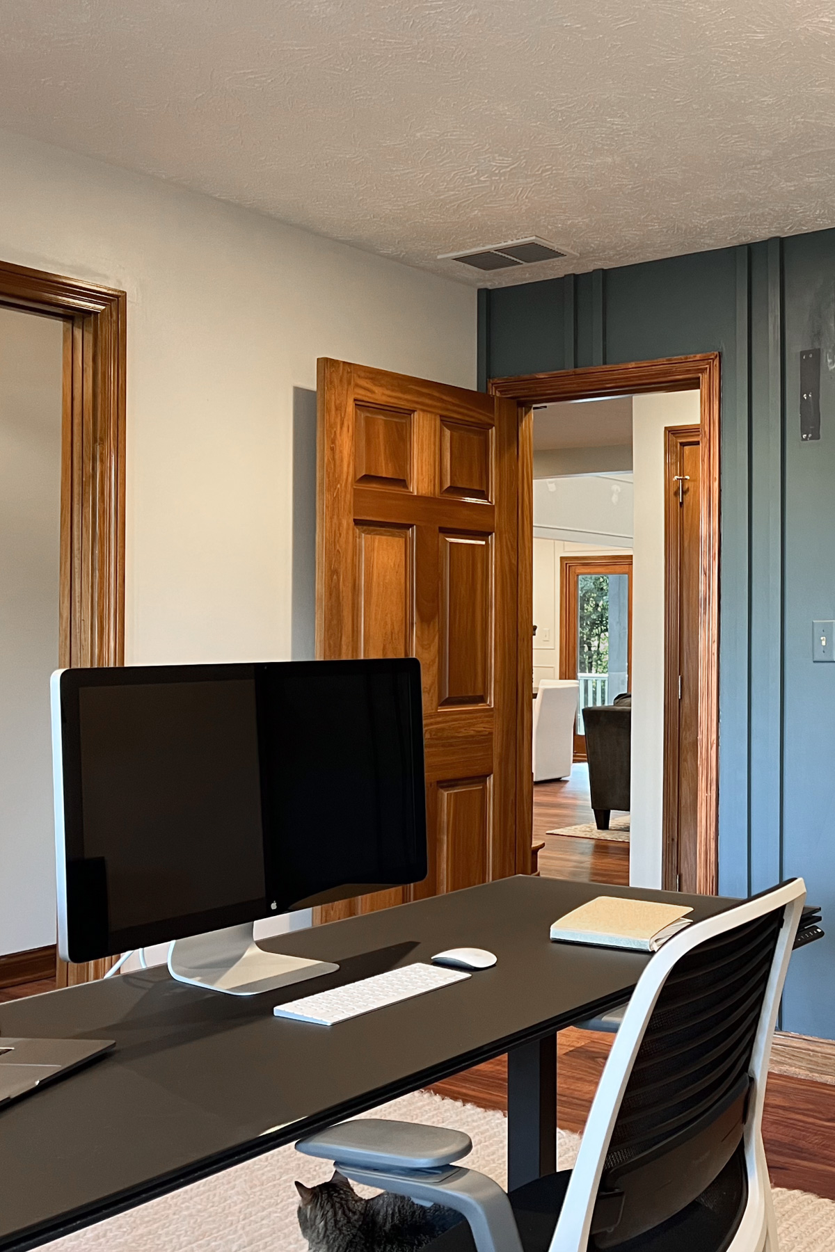 A home office featuring a black standing desk with an Apple monitor, a black and white ergonomic chair, and a view into an adjacent living area through an open wooden door.