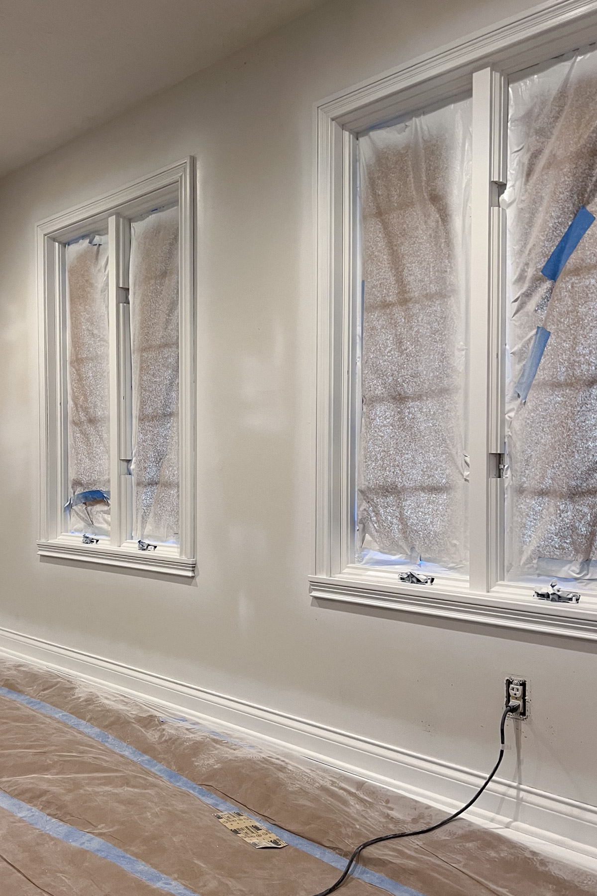 An interior wall being painted white, featuring two windows and floor sections covered in protective plastic and brown paper for renovation.