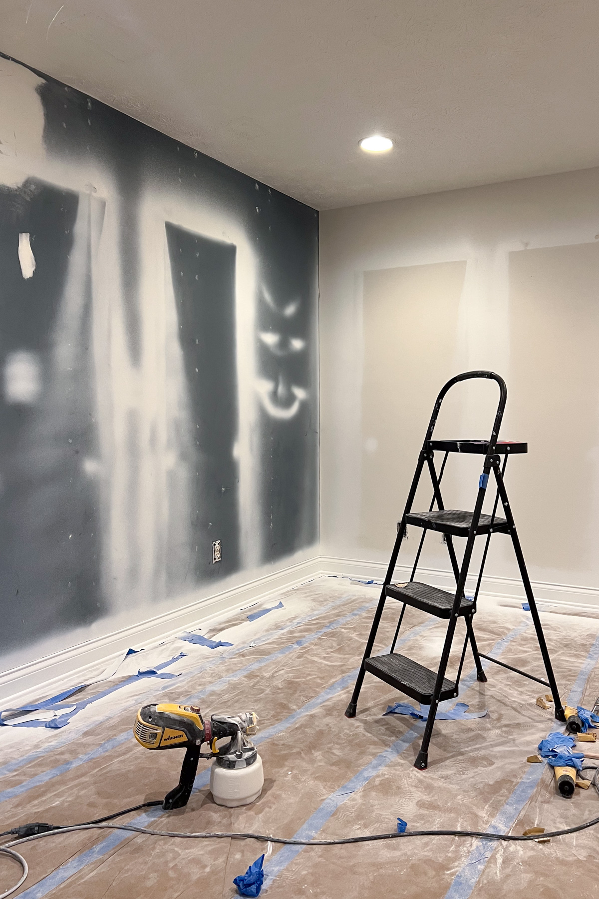 An interior room under renovation with a black step ladder, a paint sprayer on a paper-covered floor, and walls featuring navy blue and white paint tests.