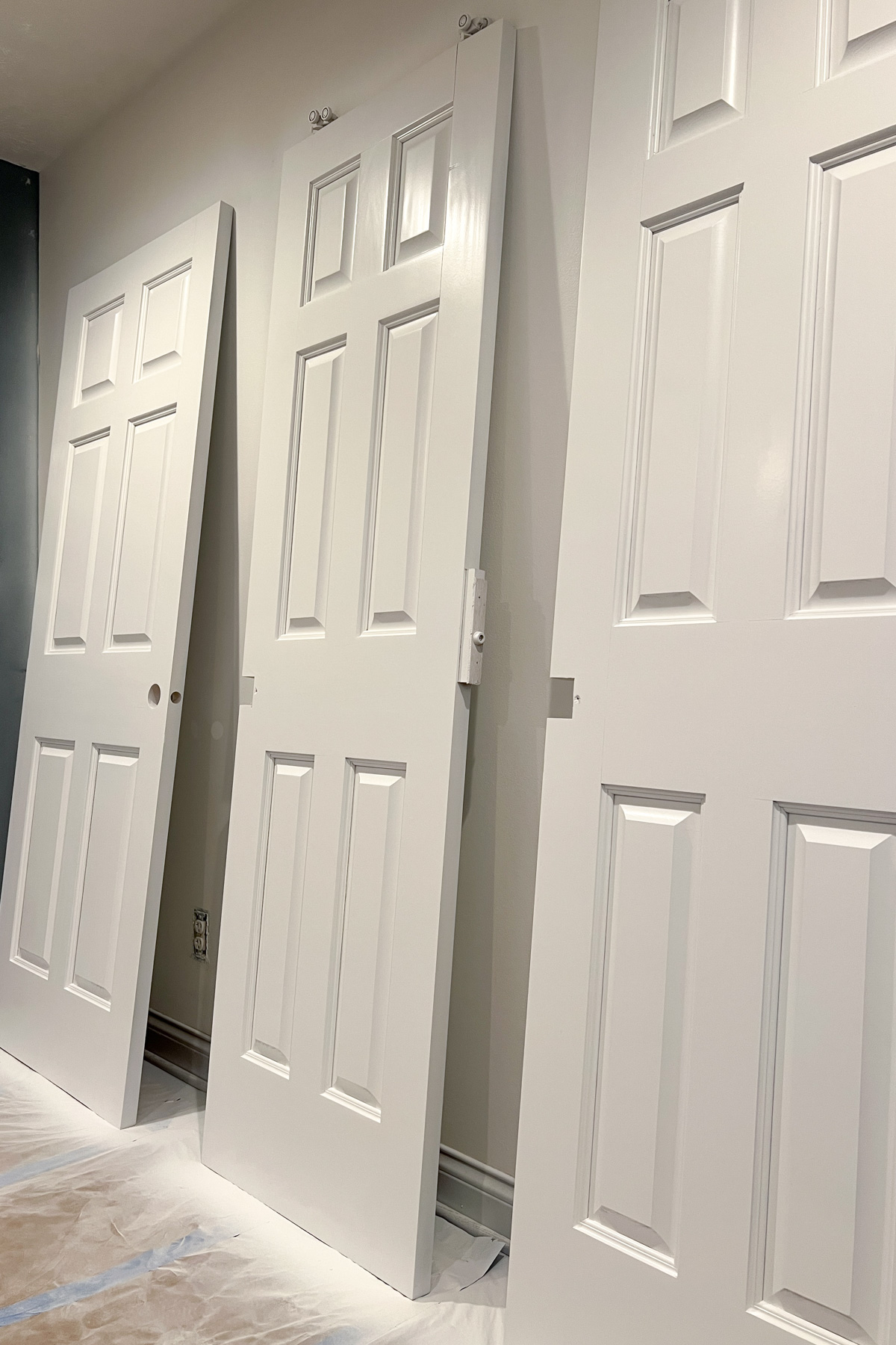 Three classic six-panel interior doors painted white, leaning against a neutral-colored wall on a floor protected by white plastic and brown paper.