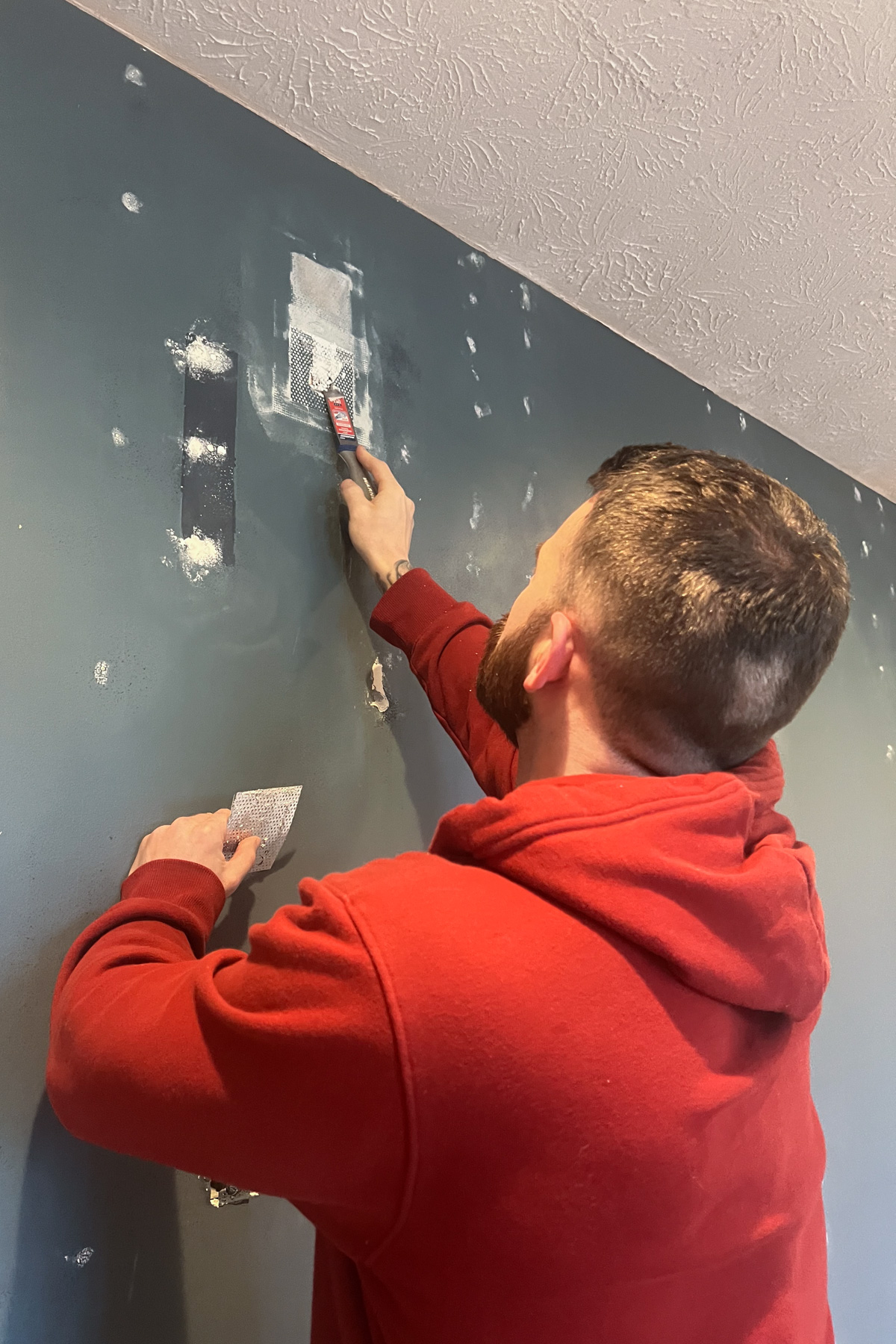 A person in a red hoodie using a putty knife to apply spackle over a mesh patch on a navy blue wall.