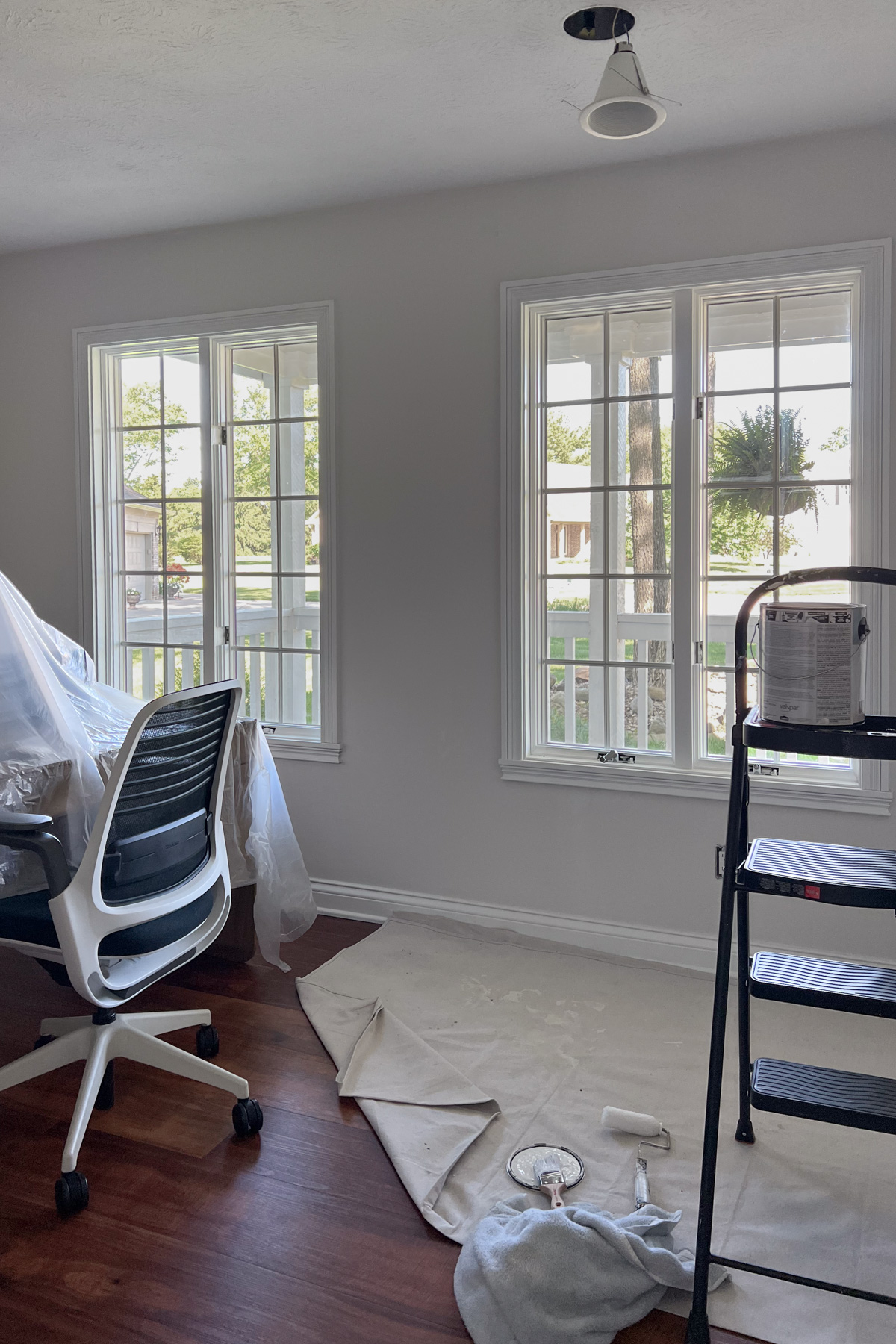 A bright room with two large windows, a black and white office chair, and painting supplies including a paint can on a ladder and a roller and brush on a drop cloth.