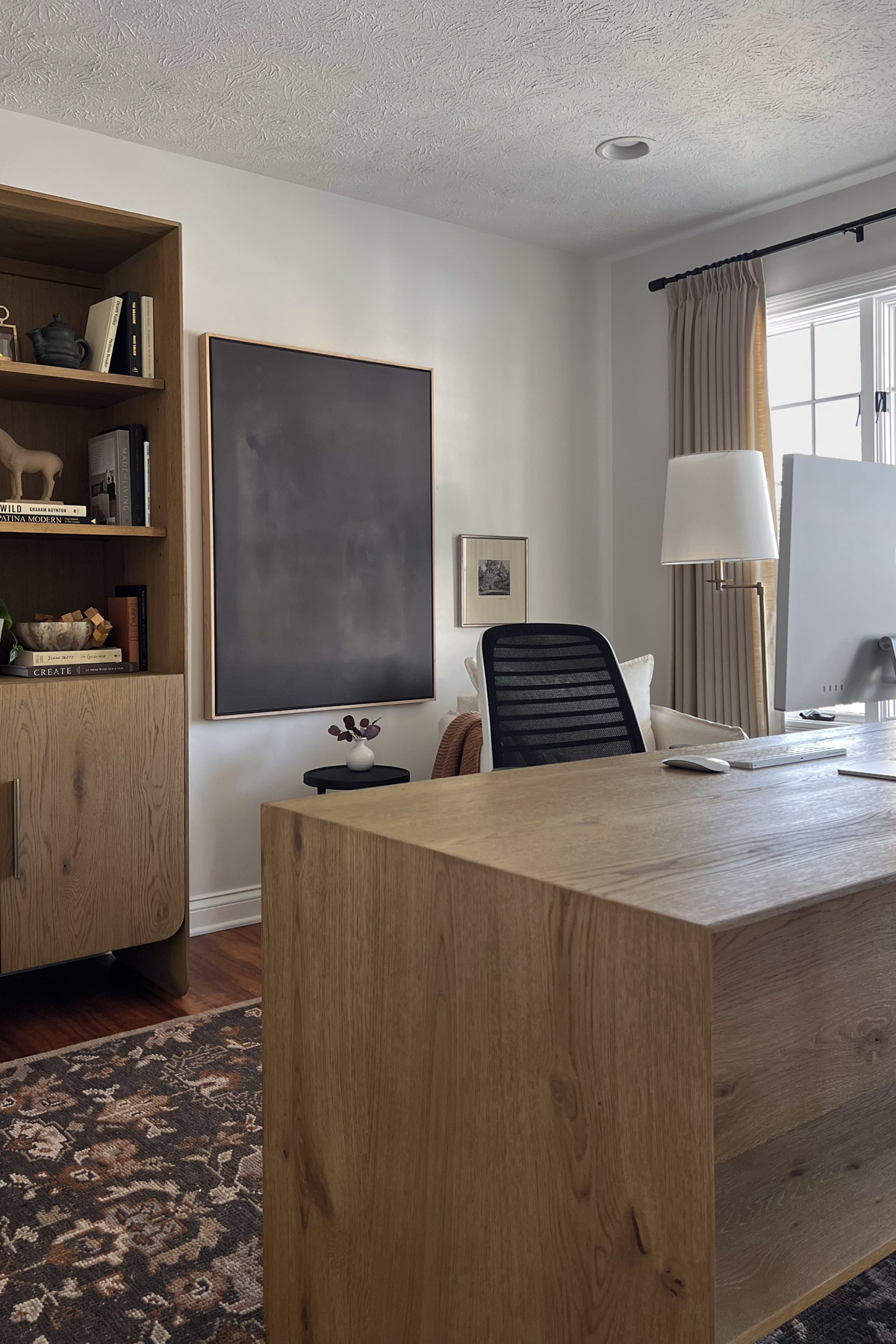A completed home office featuring a large wooden desk, a matching bookshelf, a modern office chair, and neutral-toned curtains by a window.
