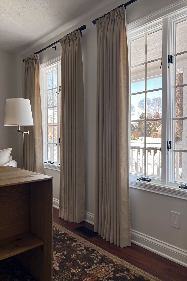Two large windows with long taupe curtains hanging from a black rod, overlooking a snowy outdoor scene.