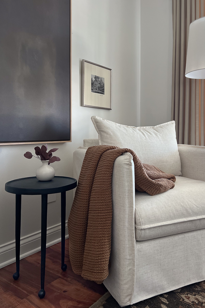 A close-up of a cream-colored armchair with a textured throw blanket next to a black pedestal side table.