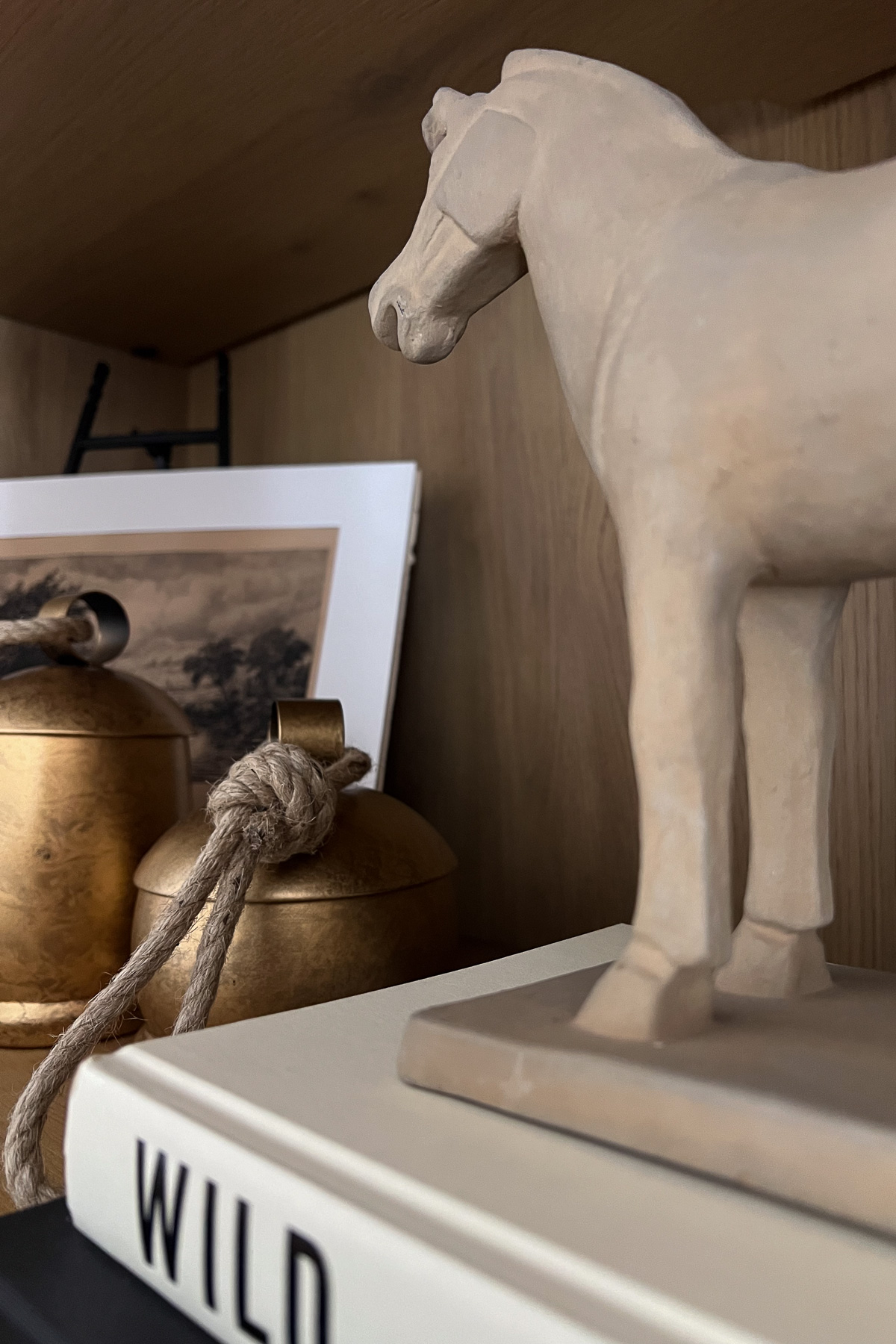A close-up shot focusing on the clay horse figurine standing on a book titled "WILD," with brass bells and a framed sketch in the background.