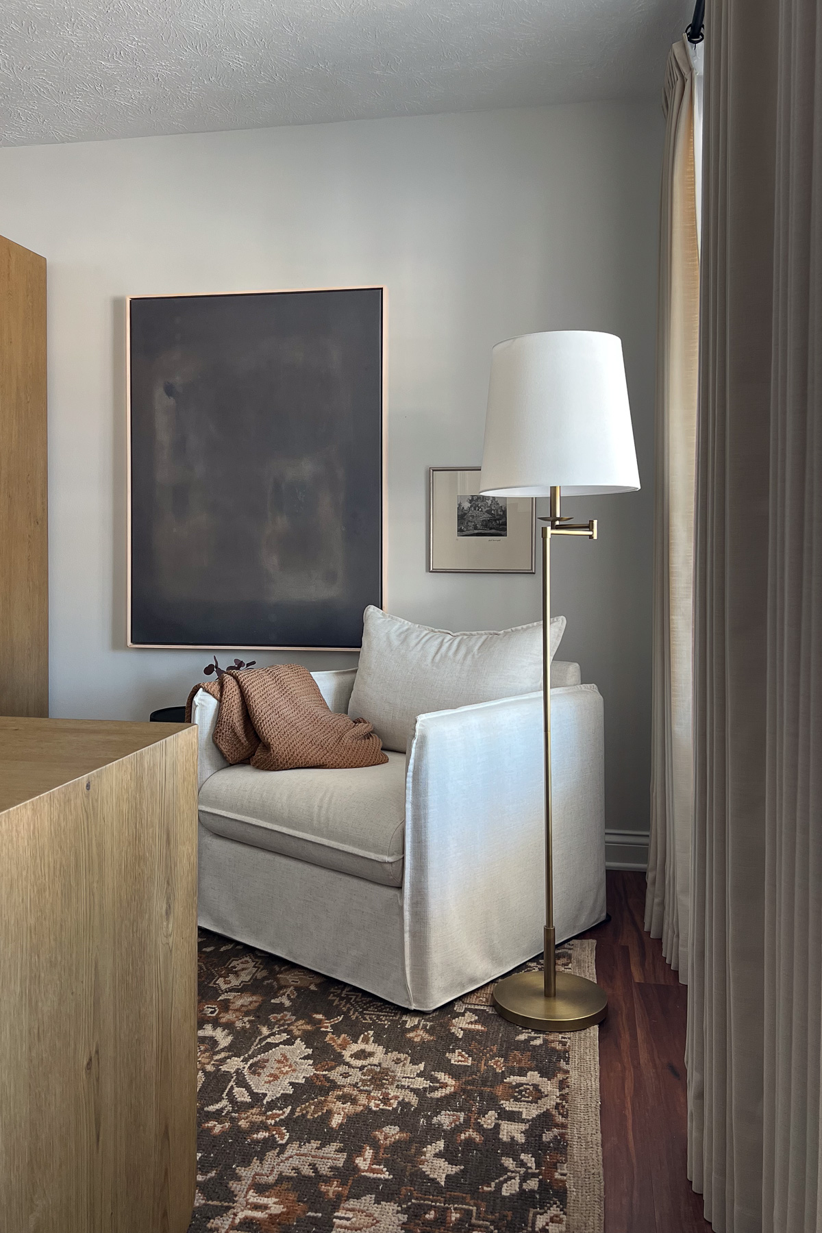 cozy, decorated corner of a room featuring a cream-colored armchair with a rust colored throw, a brass floor lamp, and a large dark abstract painting on a light wall.