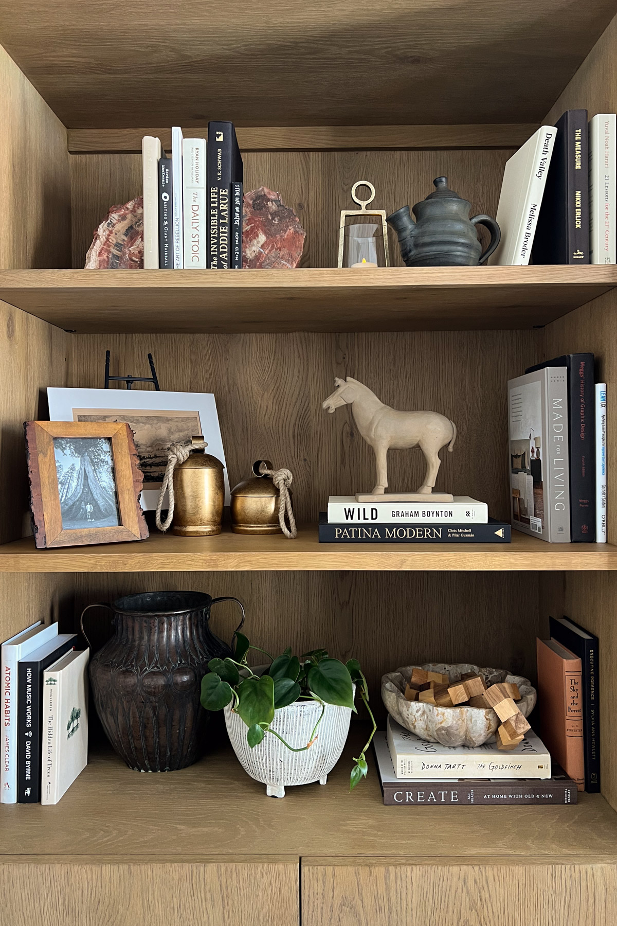 A fully styled wooden bookshelf featuring various books, a potted plant, a clay horse figurine, and decorative items like a dark ceramic teapot and a large textured metal vase.
