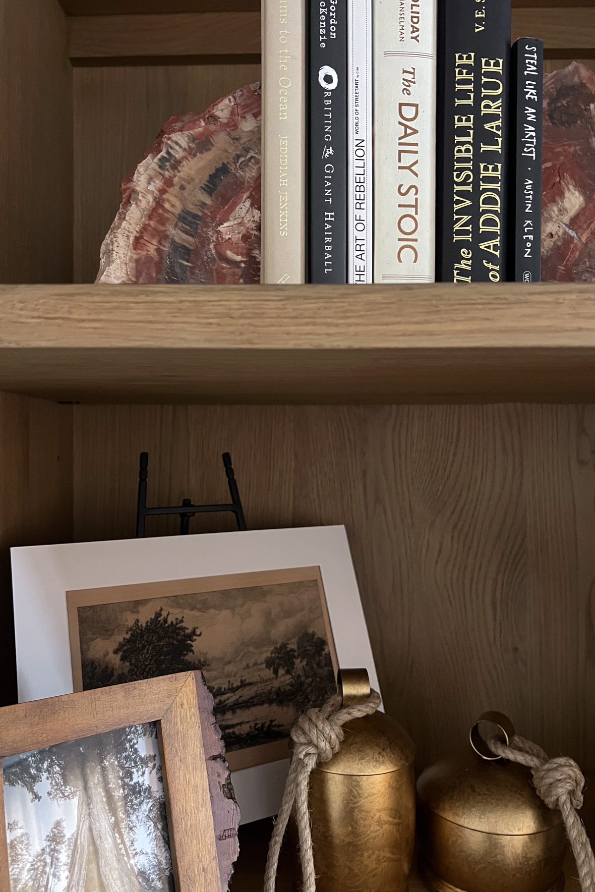 A close-up of a wooden bookshelf displaying several books including "The Daily Stoic" and "The Invisible Life of Addie LaRue," alongside decorative items like a landscape sketch and gold-toned containers.