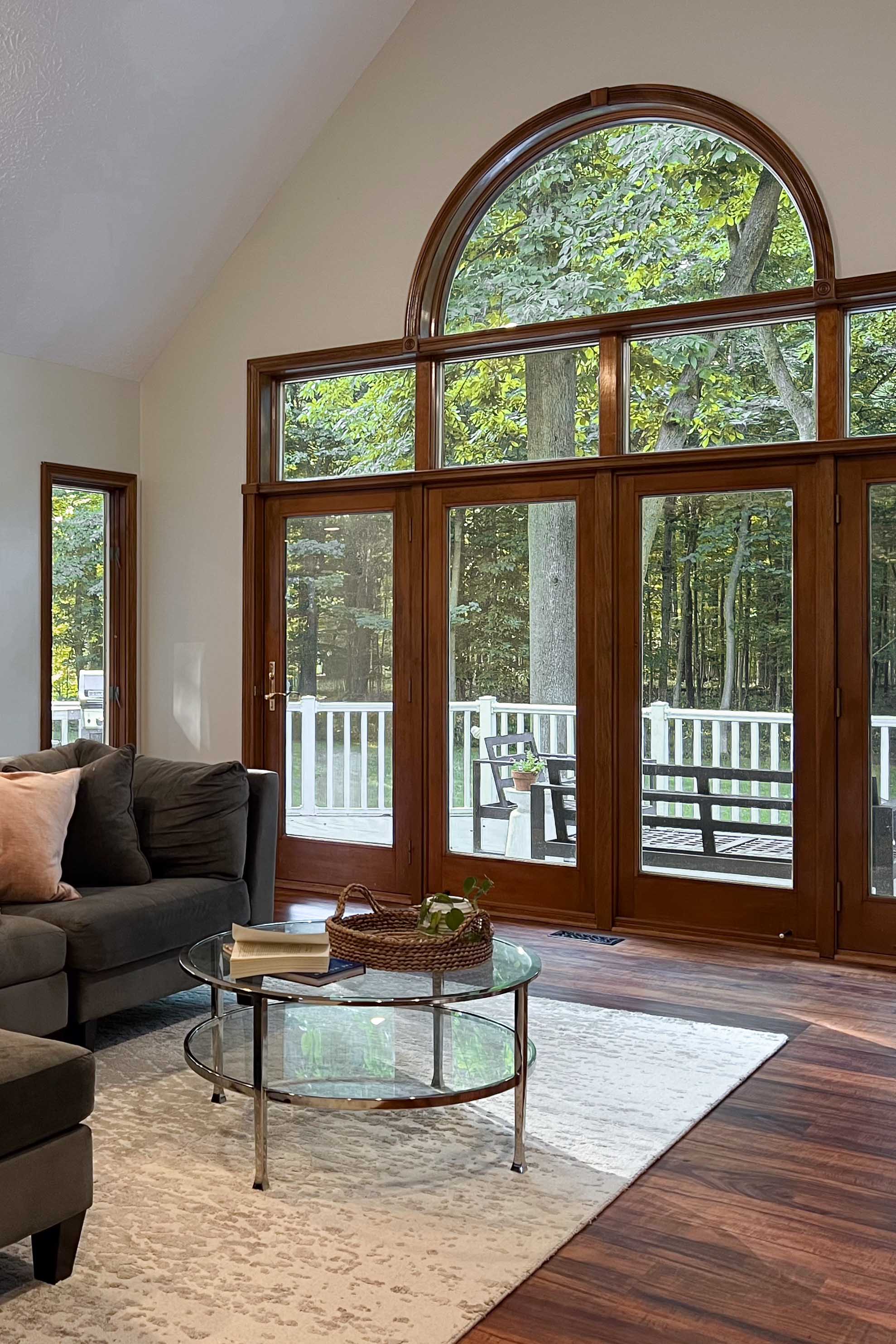 A bright living room with large wood-framed glass patio doors topped by an arched transom window, offering a view of a lush green forest and outdoor deck.