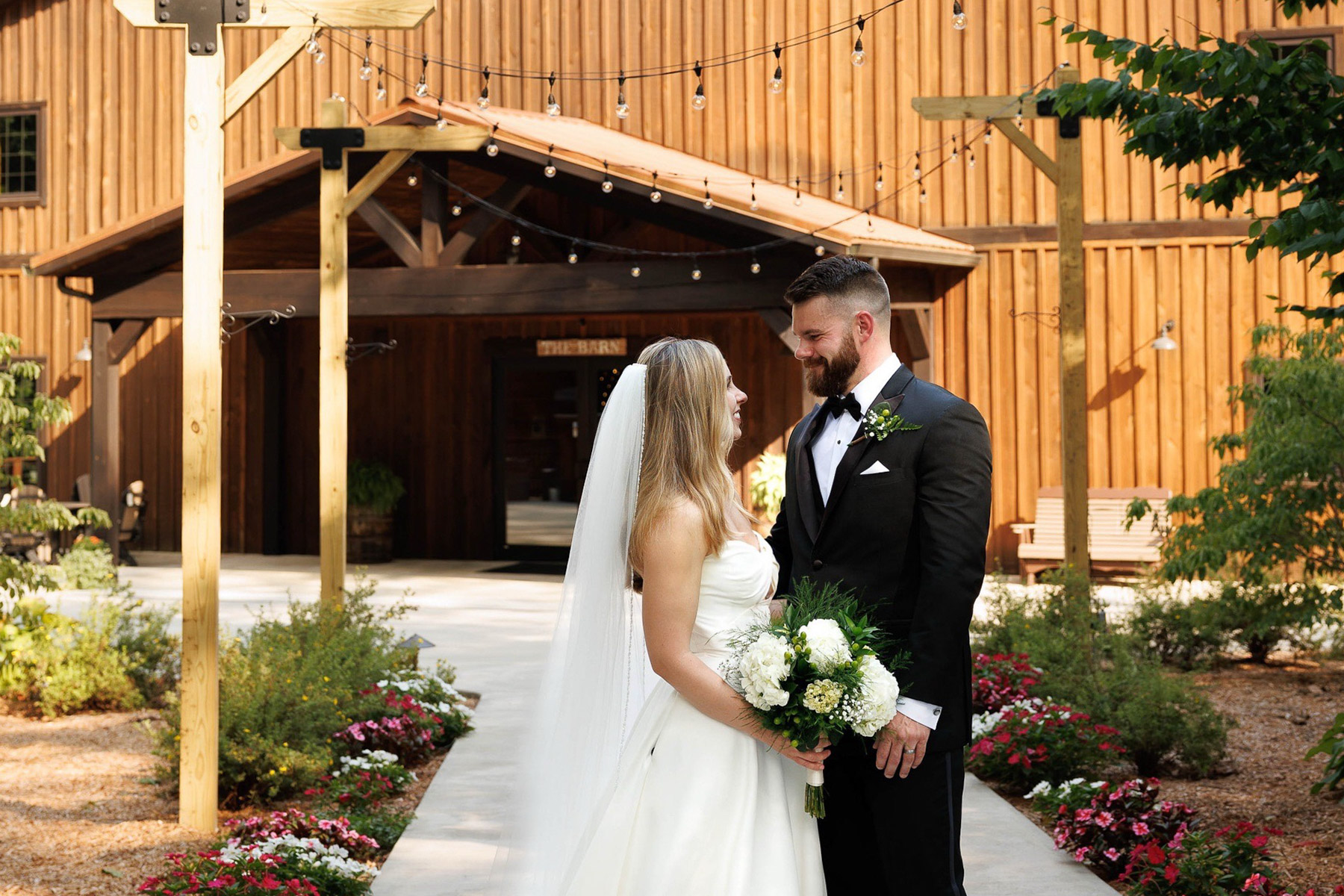 A bride in a white gown with a long veil and groom in a black tuxedo holding a white and green bouquet look at each other outside a rustic wooden barn venue with string lights.