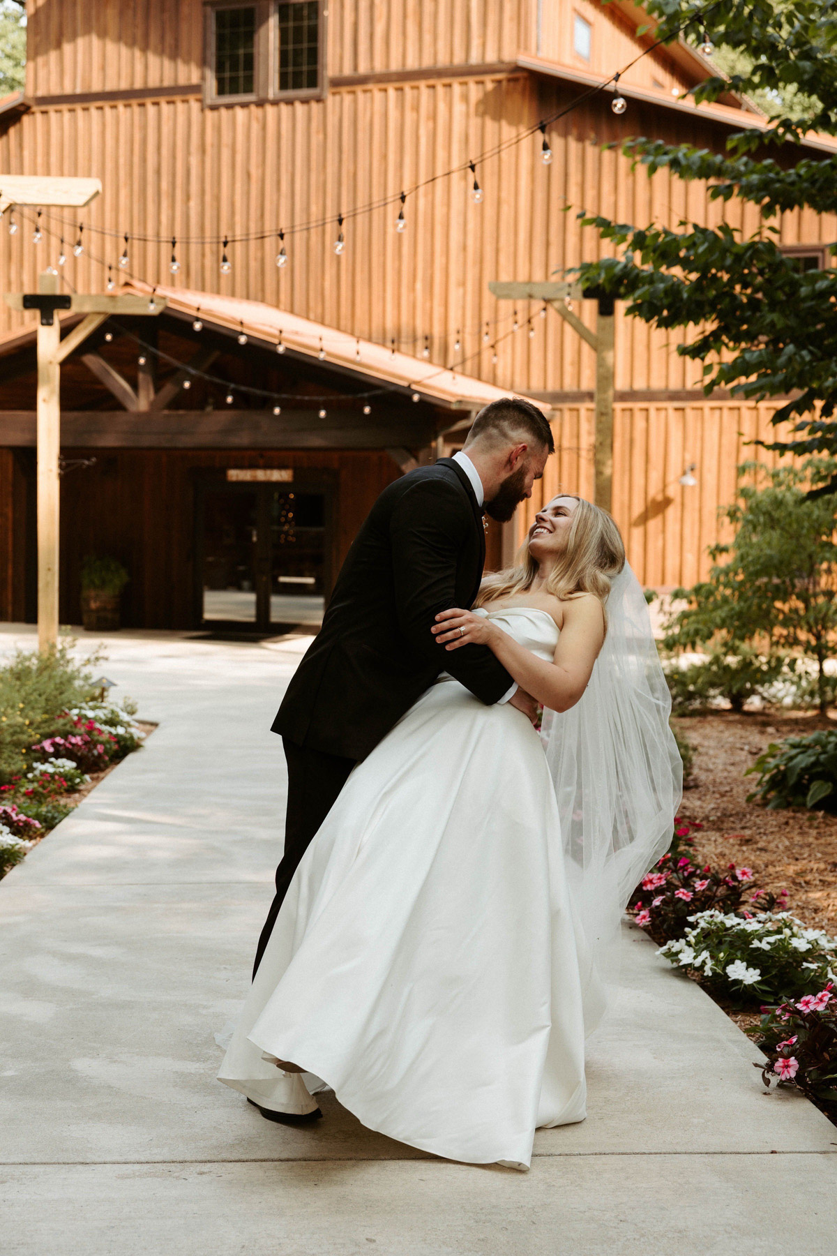 A groom in a black tuxedo dips a smiling bride in a white strapless gown on a outdoor paved walkway in front of a wooden barn venue.
