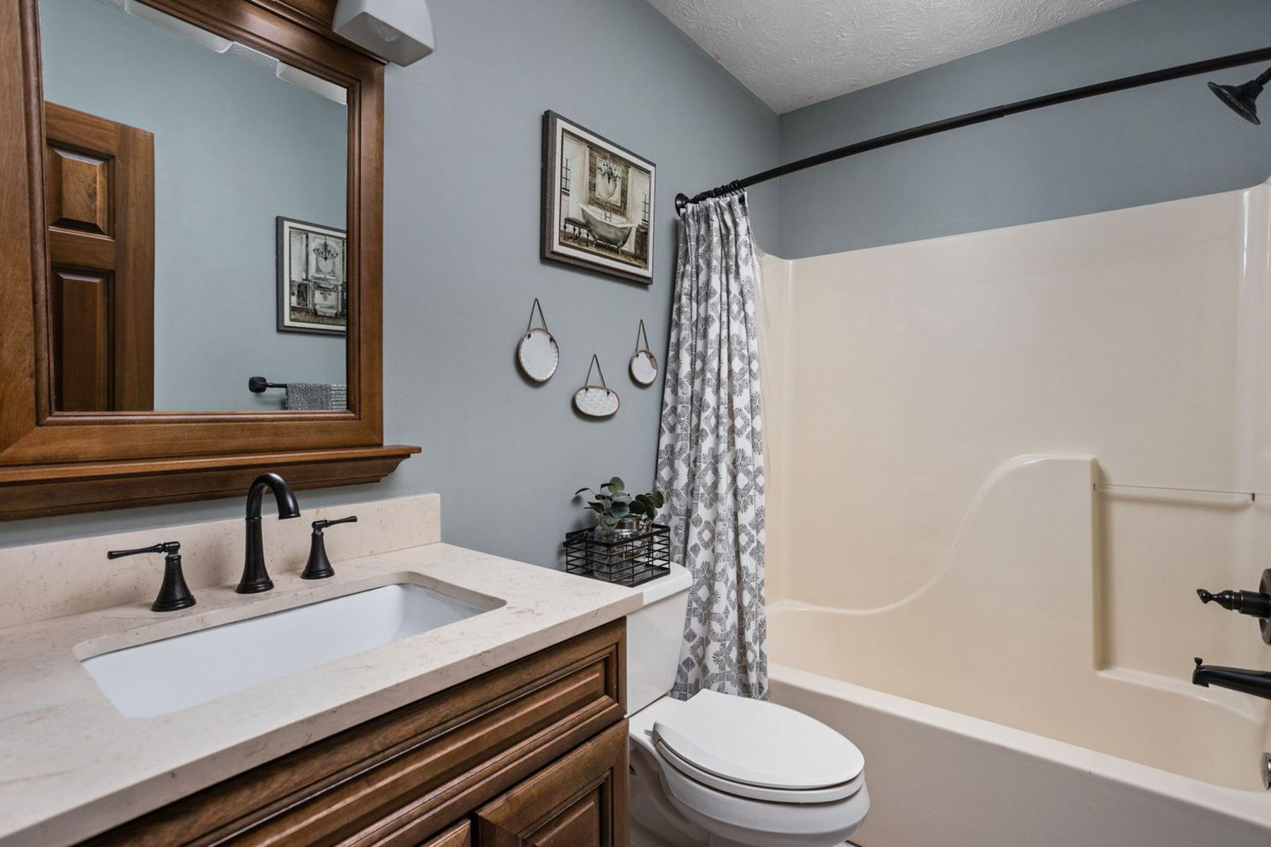 A bathroom featuring slate blue walls, a wood vanity with a light countertop, a white toilet, framed wall decor, and a white shower-tub combo with a patterned curtain.