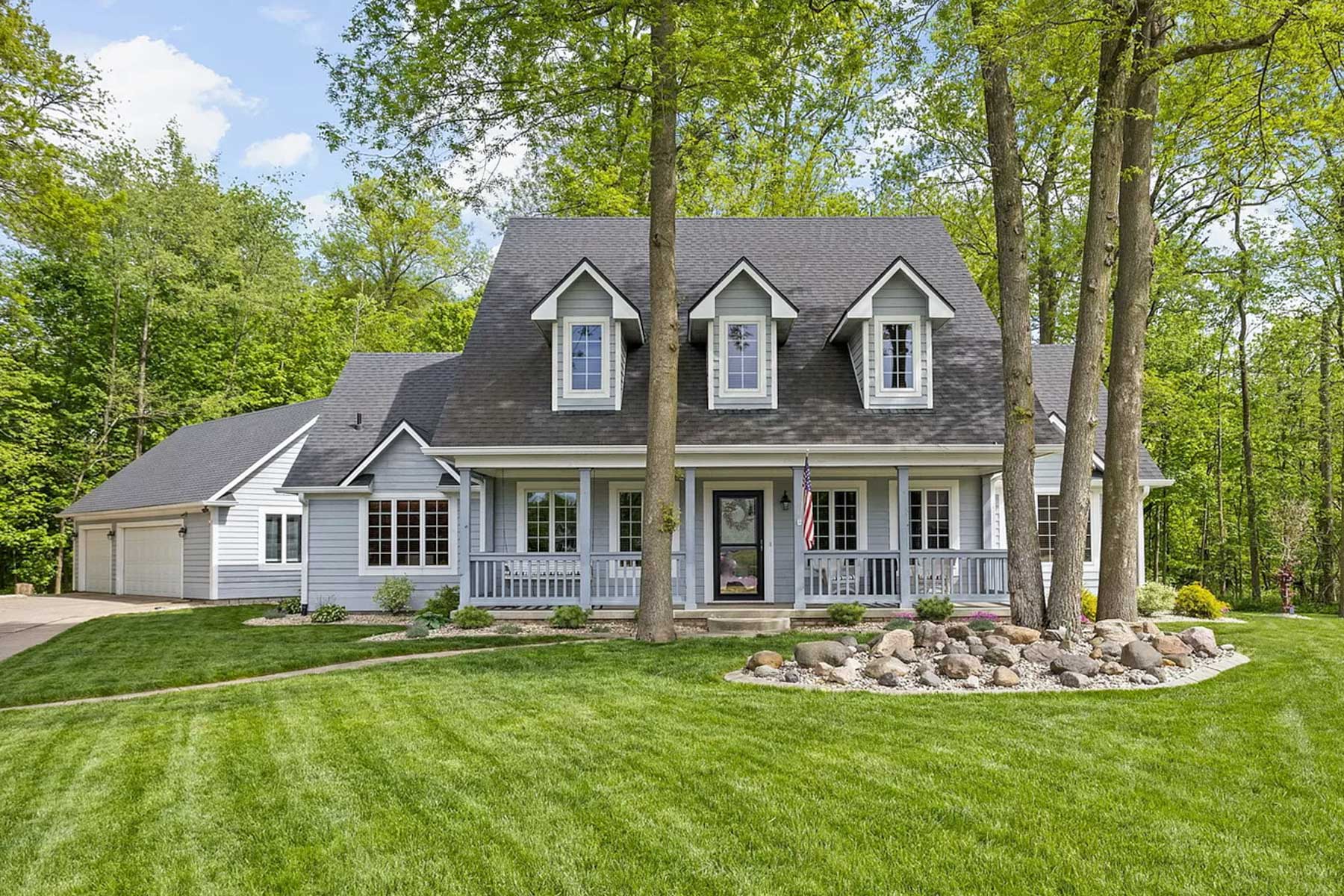 Front view of a light blue two-story house featuring three dormer windows, a full-length front porch with white railings, a dark roof, attached garage, and a landscaped rock bed surrounded by mature trees on a sloped green lawn.