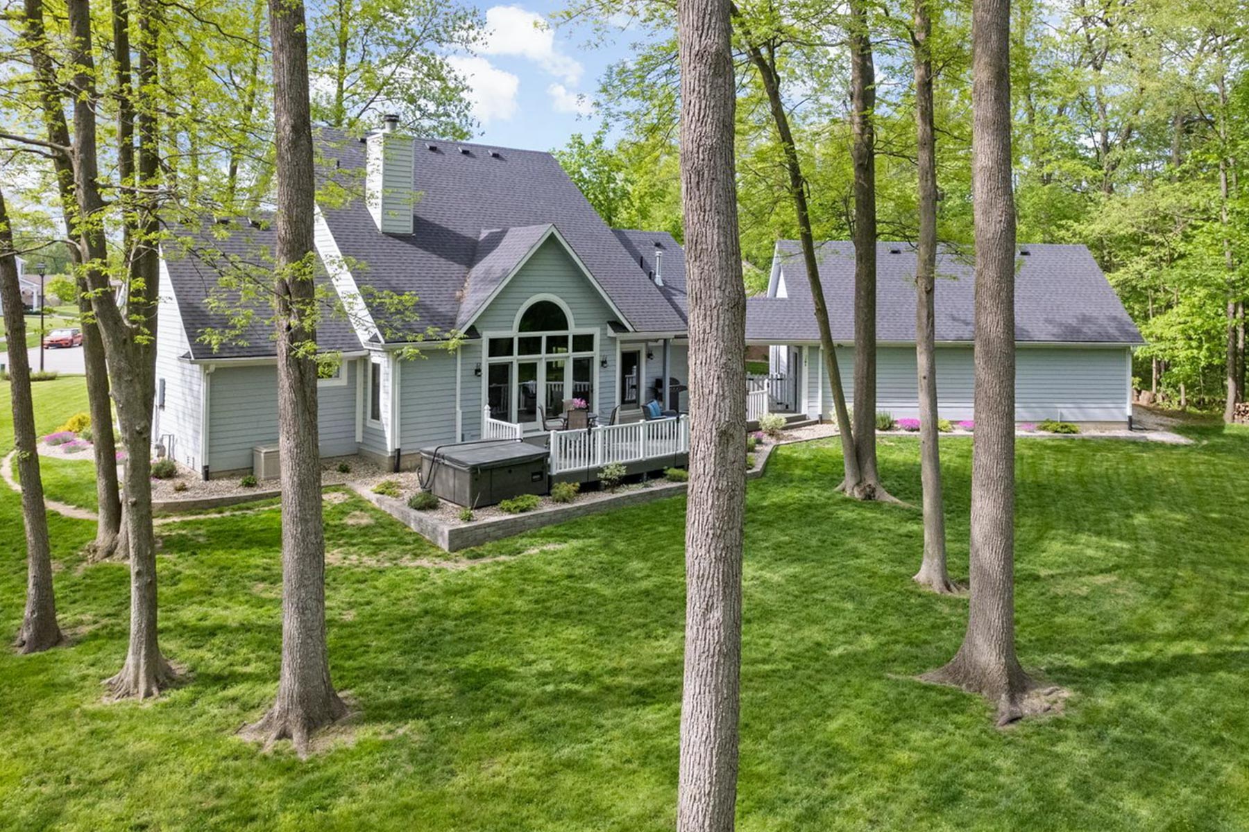 Rear view of the home showing a elevated back deck with white railings, a hot tub, large arched window, a detached or breezeway-connected garage, and a grassy backyard shaded by tall trees.
