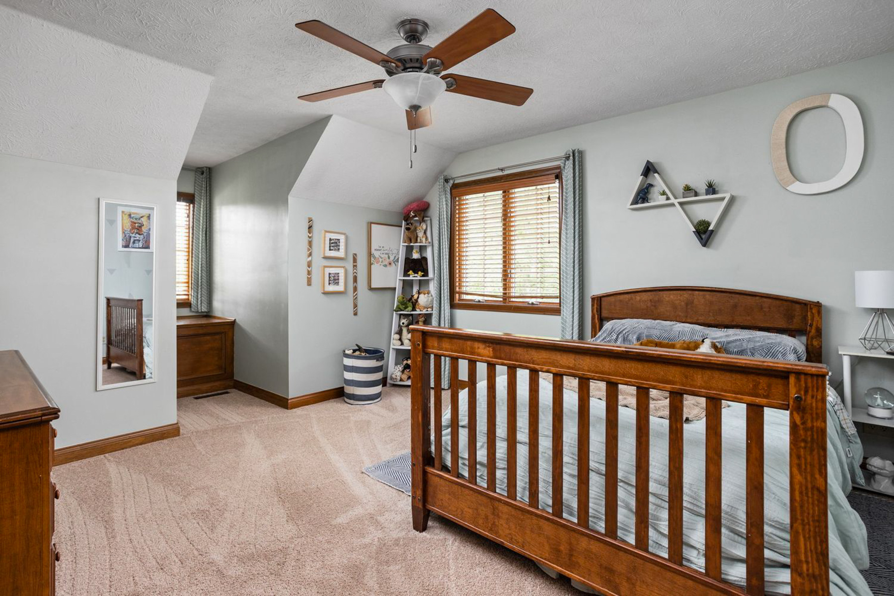 View of a bedroom with light mint green walls, beige carpeting, and warm oak wood trim. A wooden slatted bed with light blue bedding sits in the center. The wall behind the bed is decorated with a geometric shelf, a large white letter "O", framed art, and a window with wooden blinds and curtains. A window nook with a built-in wooden cabinet is visible in the background.