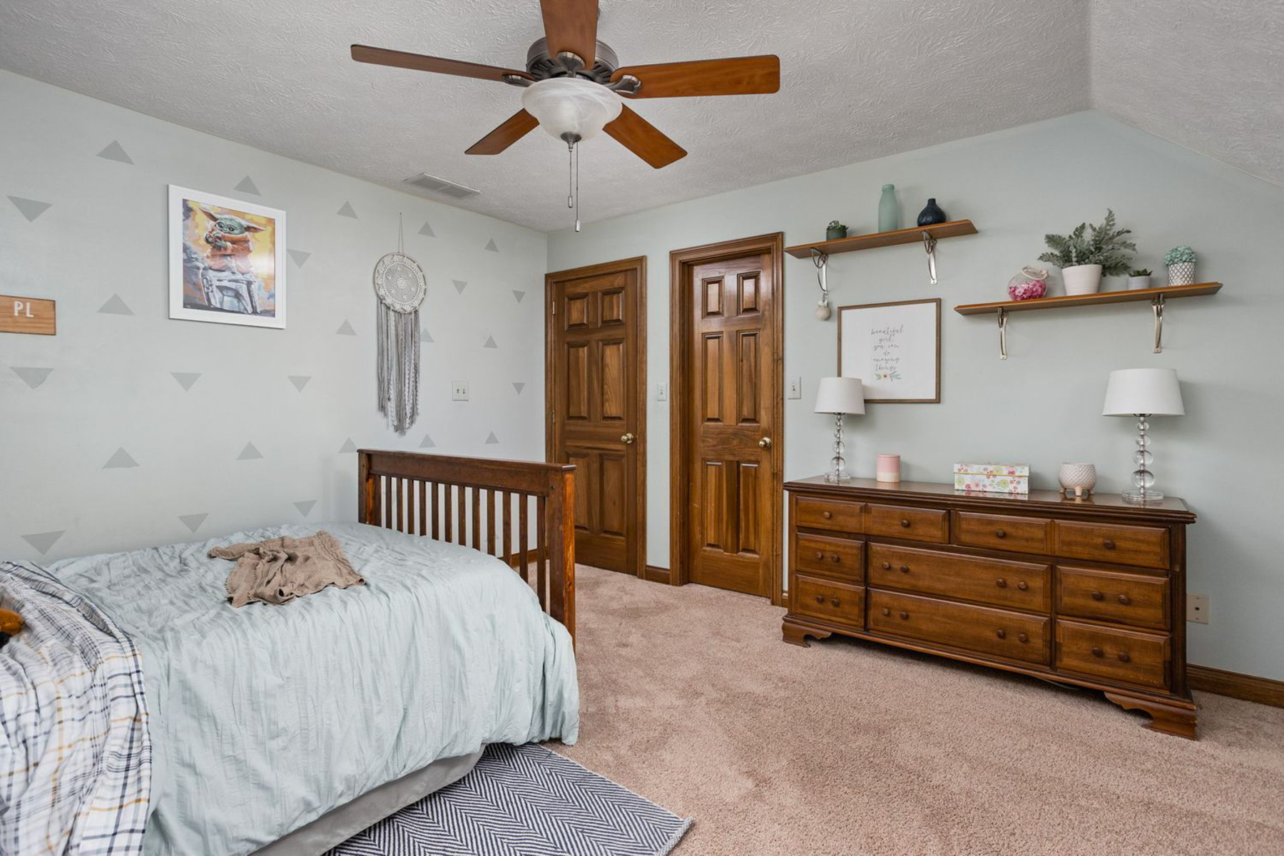 View showing the opposite side of the bedroom with light mint green walls painted with a subtle blue triangle pattern. A wooden bed sits in the foreground beside two panel doors with warm wood trim. Along the right wall, a long wooden dresser sits beneath floating shelves decorated with plants and artwork, and a dreamcatcher hangs on the wall near a framed print.