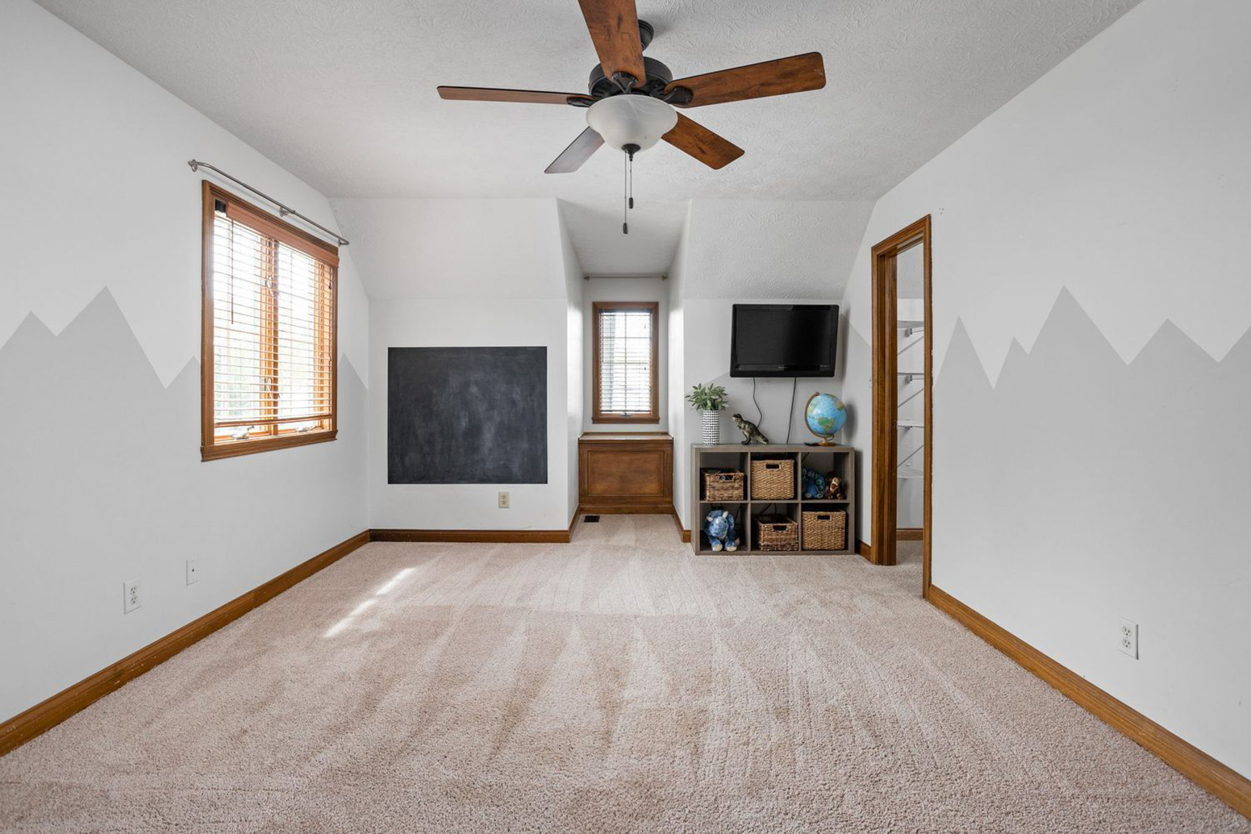 An empty upstairs bedroom or playroom with light gray mountain mural walls, wood trim, beige carpet, a ceiling fan, a wall-mounted chalkboard, and a cubby organizer with a TV and globe.
