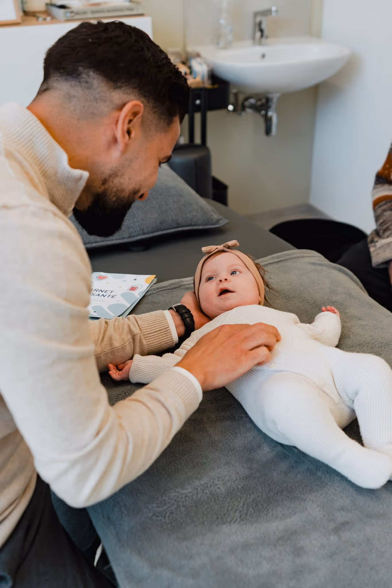 Man smiling and gently touching a baby dressed in white lying on a gray blanket.