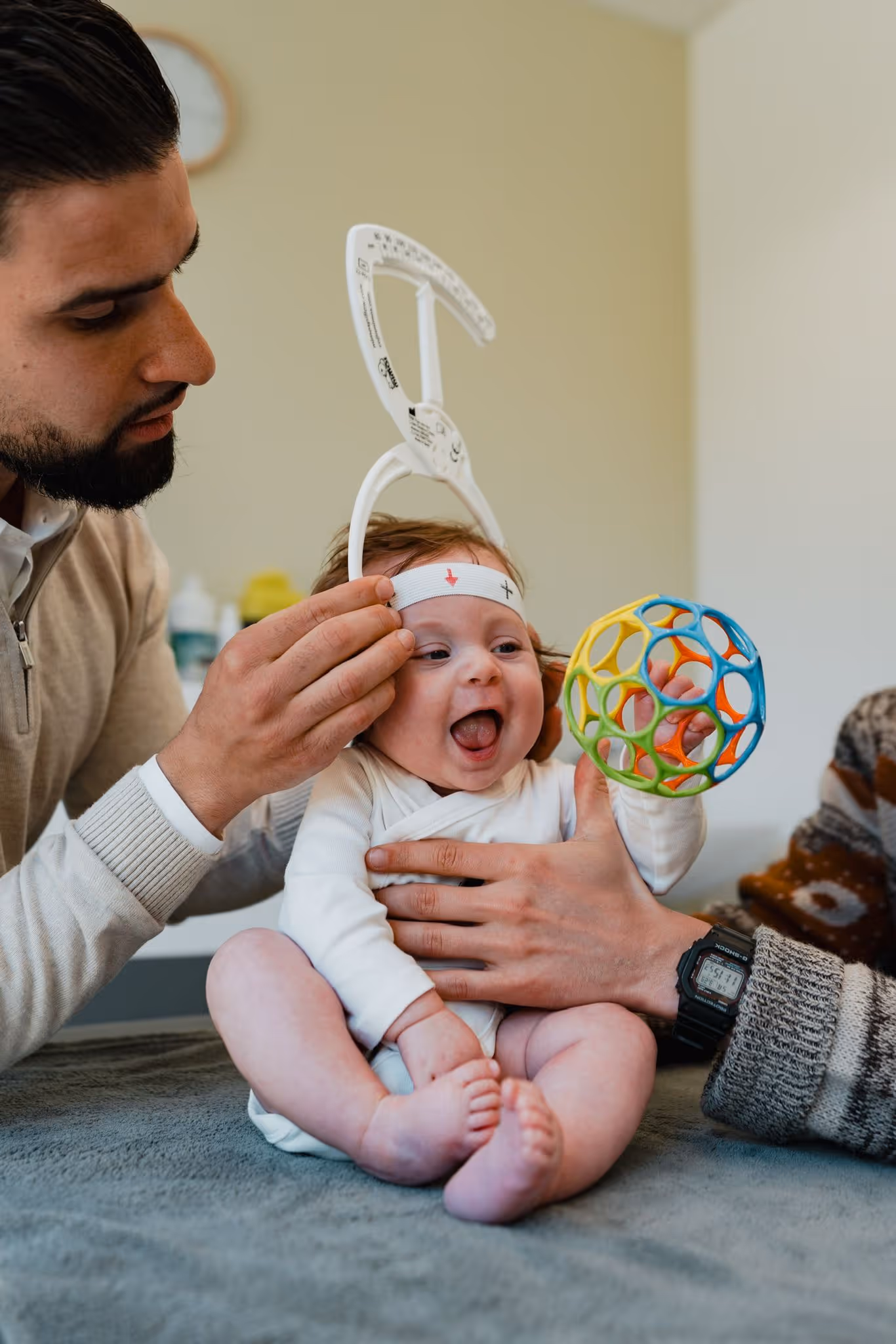 A man gently measures a smiling baby’s head circumference with a white measuring tape while another person holds a colorful toy ball.