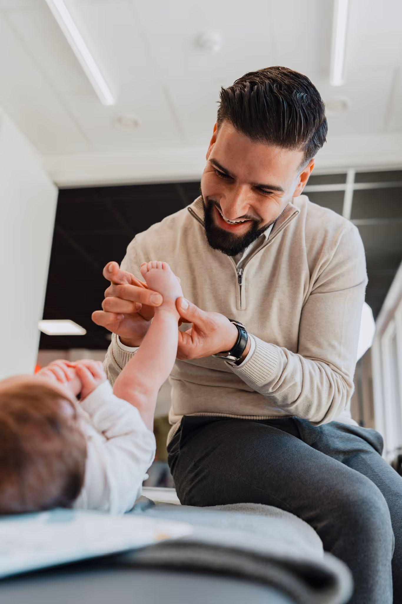 Smiling man gently holding and playing with a baby's foot while sitting indoors.