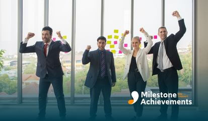 A diverse business team in formal attire celebrating success with raised fists in front of a large window.