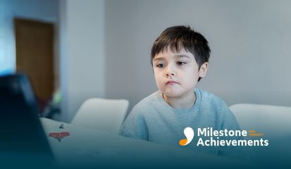A young autistic child sitting at a table, staring intently at a laptop screen, with scissors and paper nearby.