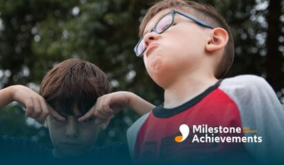 Two boys stand outside making silly faces, one wearing glasses, both bursting with playful energy and carefree joy.