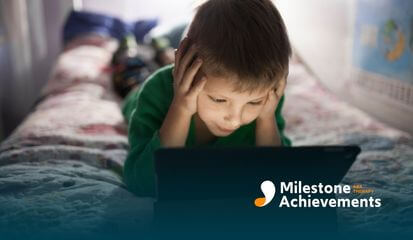 A boy sits on his bed, holding his head in frustration, feeling overwhelmed as he struggles with the tablet in front of him.