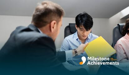 Two people in a business setting, one presenting a yellow folder to the other, with others seated around a table.