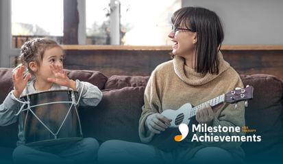 Mother and young child play music together on a couch, smiling as the child drums and the parent strums a ukulele at home.
