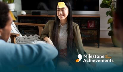 Young woman sits on the floor during a group activity with a note on her forehead, smiling and engaging with others.