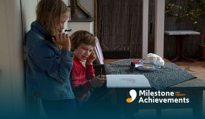 Two kids at a table, one covering ears while drawing, the other holding an object, showing sensory discomfort.