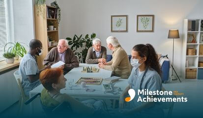 Caregivers and older adults gathered at table in group home, playing games and talking in supportive shared living space.