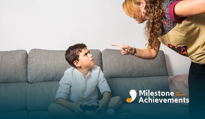Adult points while speaking to a child holding a game controller on a couch, illustrating a challenging behavior moment.