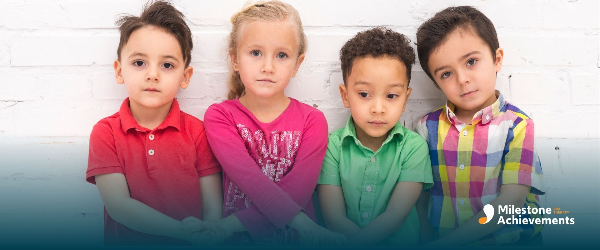 Four autistic children, standing side by side, holding hands in front of a white brick wall, each wearing colorful shirts.