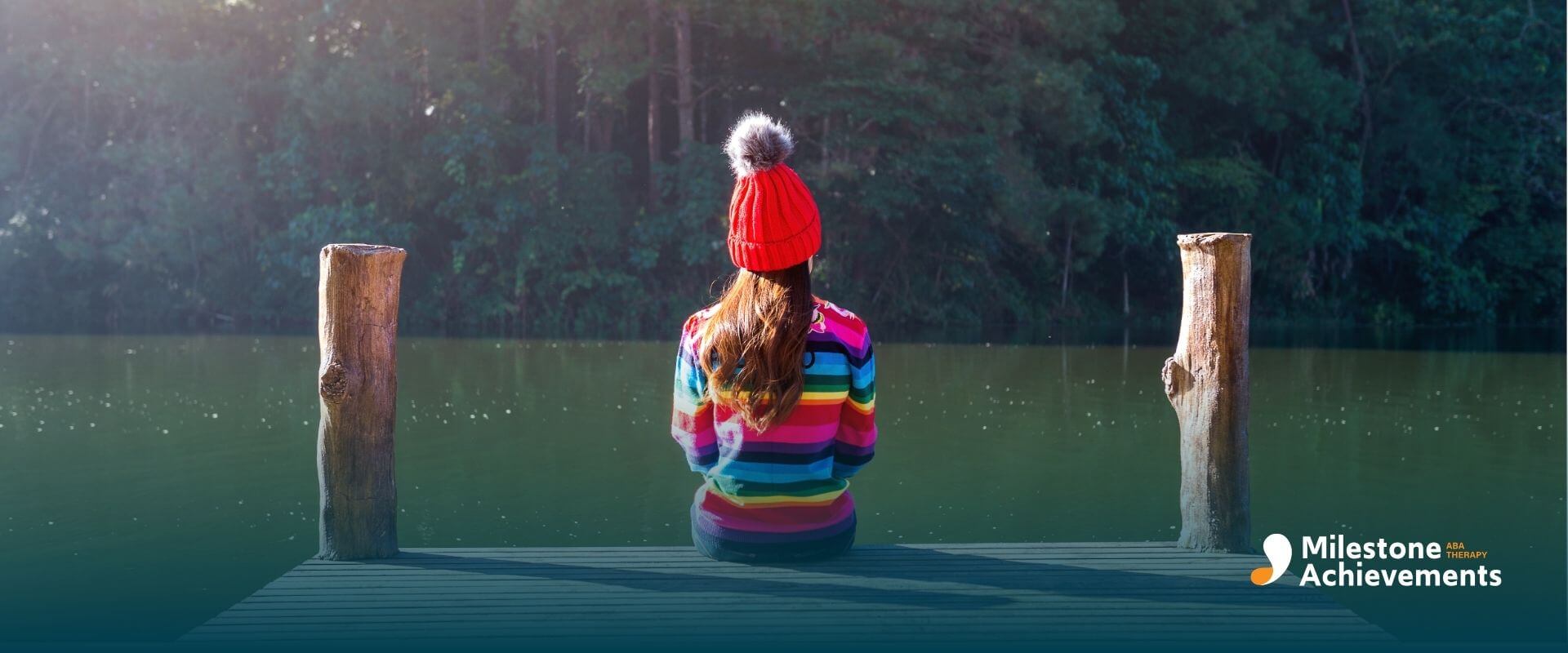 Autistic child wearing a rainbow sweater sitting alone on a wooden dock by a calm lake after ABA therapy