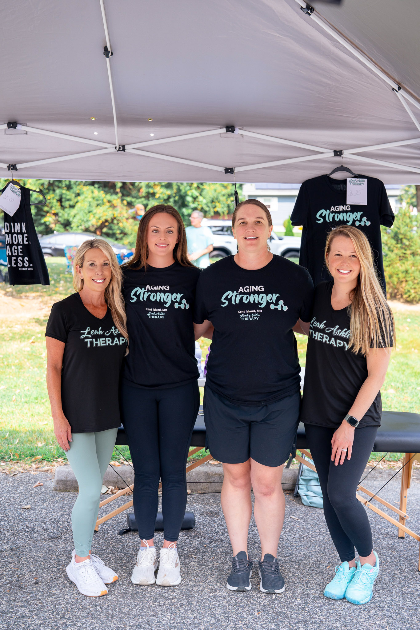 The team of a physical therapist office is standing under a tent at an outdoors event. The four of them are standing shoulder to shoulder and smiling.