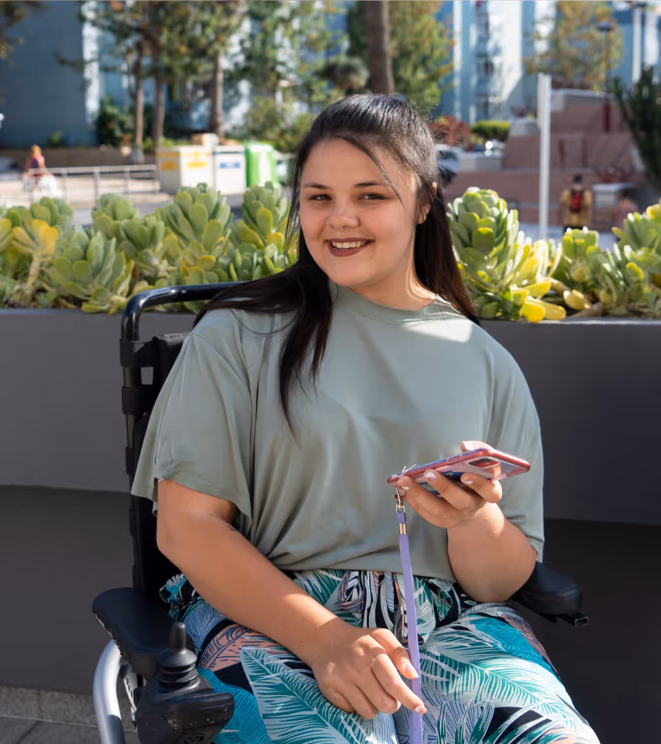 A young woman in a wheelchair smiling confidently at the  camera while holding her phone, representing inclusive  employment opportunities through GoHeadHunt.