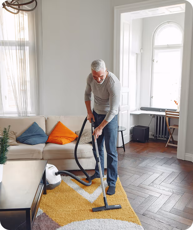 A man is vacuuming a tidy living room, focusing on the carpet while surrounded by furniture and decor.
