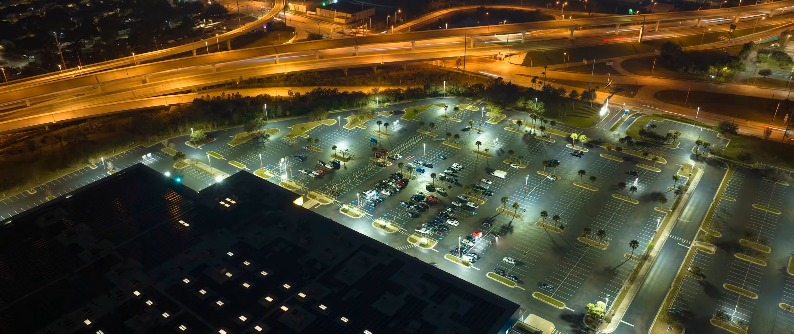 Aerial night view of a large, lit commercial parking lot with a highway network in the background.