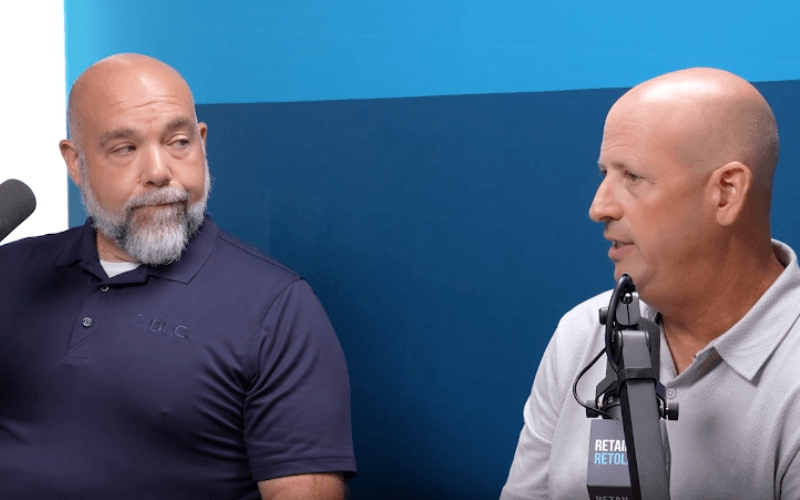 Two men sitting side-by-side in a podcast studio with microphones, speaking in front of a blue backdrop.