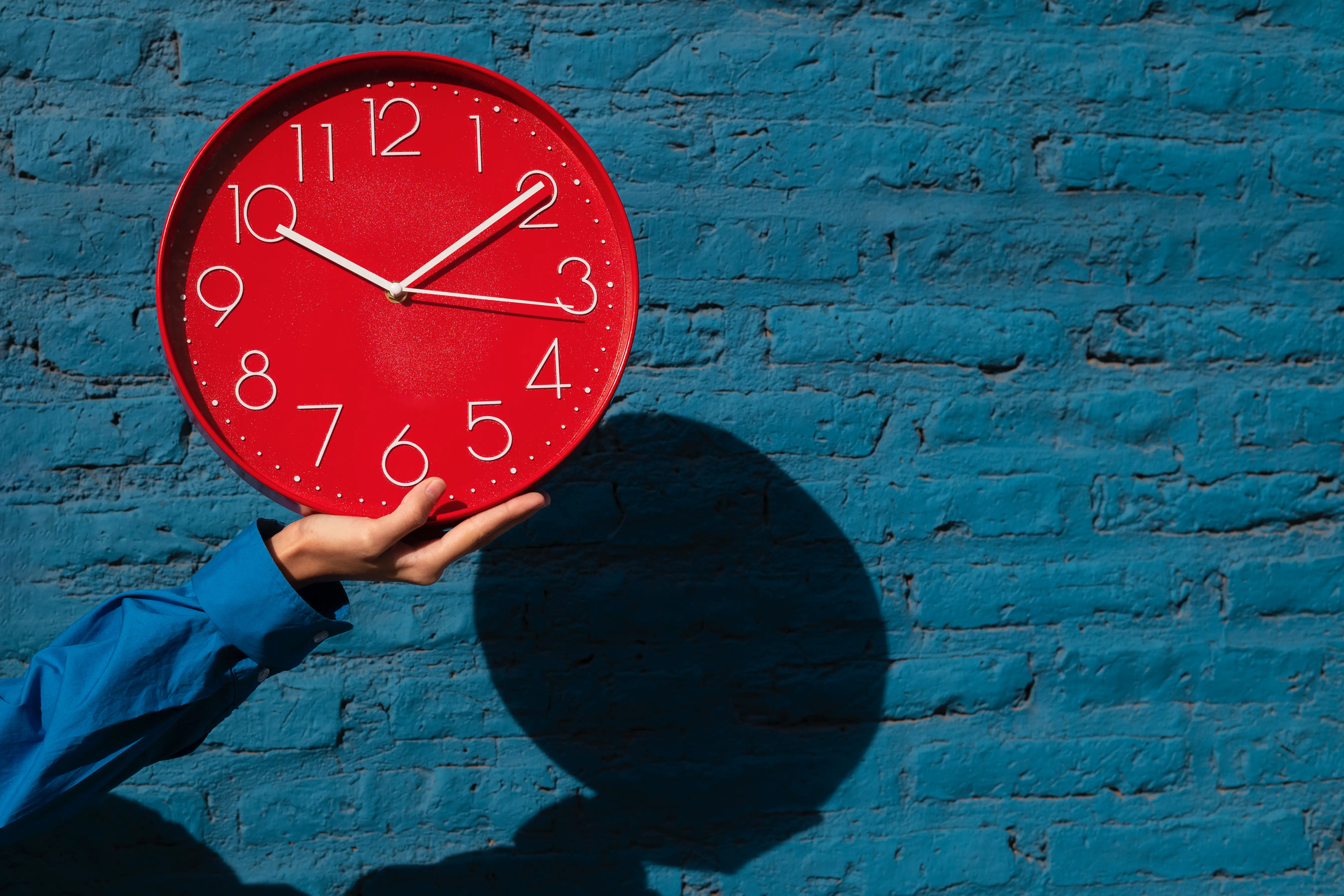 Une personne portant une chemise bleue tient une grande horloge murale rouge vif dans la paume de sa main. L'horloge se détache sur un mur de briques bleu texturé, avec une ombre portée circulaire marquée sur la droite.