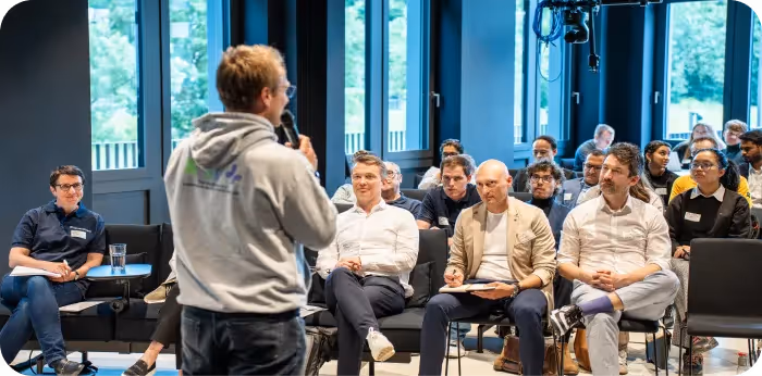 A speaker in a grey hoodie addresses an audience sitting in a bright conference room with large windows.