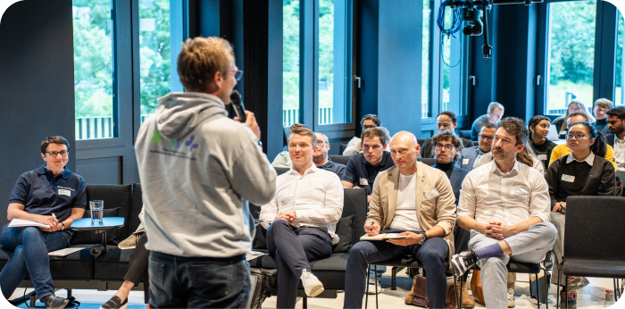 A speaker in a grey hoodie addresses an audience sitting in a bright conference room with large windows.